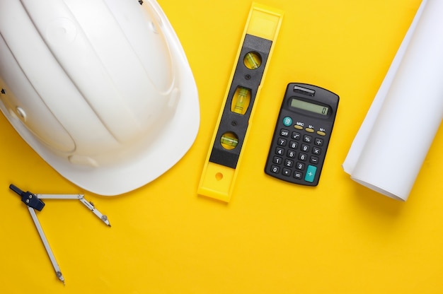 Hard hat with building level, calculator, compass, blueprint on yellow studio