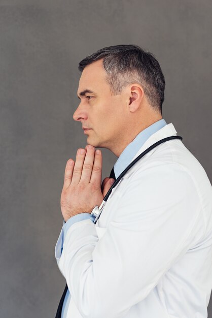 ハードメディカルケース。灰色の背景に立っている間、手を握りしめ、目をそらしている成熟した男性医師の側面図