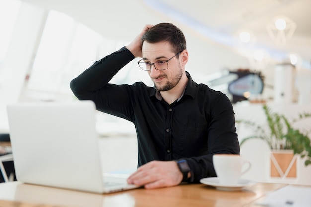 Hard remote work. Freelancer man holding his head doesn't understand what is happening in the chat with the employer. Male remote worker in black shirt working from coffeehouse and using laptop.