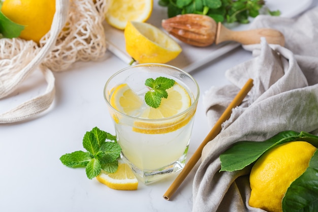 Hard seltzer cocktail with lemon, mint and ice on a table. Summer refreshing beverage, drink with trendy zero waste accessories, bamboo straw and mesh bag.