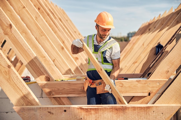 Hard-working man in a hard hat and gloves holding a piece of wood for building a roof