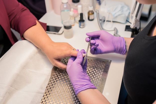 Hardware manicure in a modern beauty salon. The cosmetologist in a face pink mask does the cosmetic procedure for the blond client, the professional workplace in the background