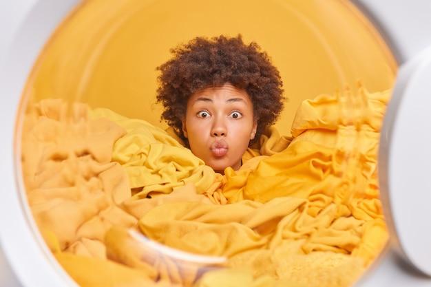 Foto hardwerkende huisvrouw met krullend haar verdronken in stapel wasgoed houdt lippen gevouwen kijkt verrassend naar voren poses van binnenkant wasmachine doet wassen