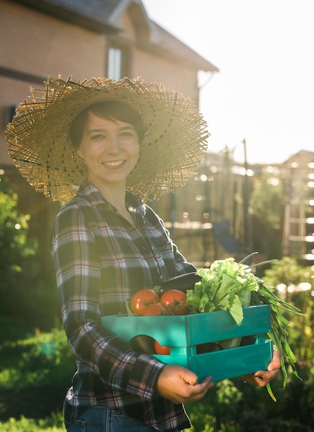 Hardwerkende jonge tuinman in strohoed pakt haar oogstdoos tomaten op zonnige zomerdag Concept van biologische landbouw en groenteteelt