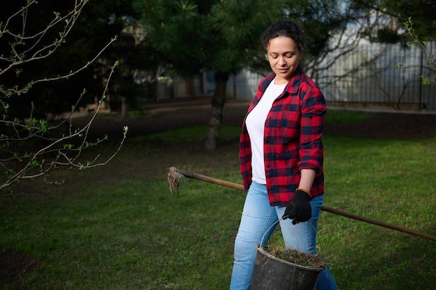 Hardwerkende vrouw tuinman werkt in de tuin houdt een emmer gemaaid gras en hooivorken en ruimt het gebied op