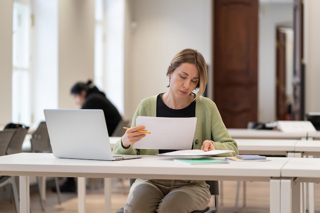 Studentessa matura laboriosa che legge disegni architettonici mentre studia nella biblioteca pubblica