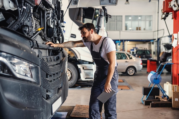 Трудолюбивый татуированный бородатый механик опирается на грузовик и проверяет двигатель, стоя в гараже импортно-экспортной фирмы.