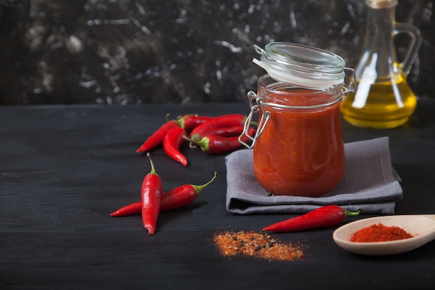 Harissa spicy herb in a glass jar stands on a gray linen napkin, a wooden spoon with spices, oil and chili pepper.