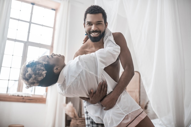 Harmonieuze relatie. jonge volwassen vrolijke donkere man shirtless en vrouw in wit overhemd dansen thuis in de middag