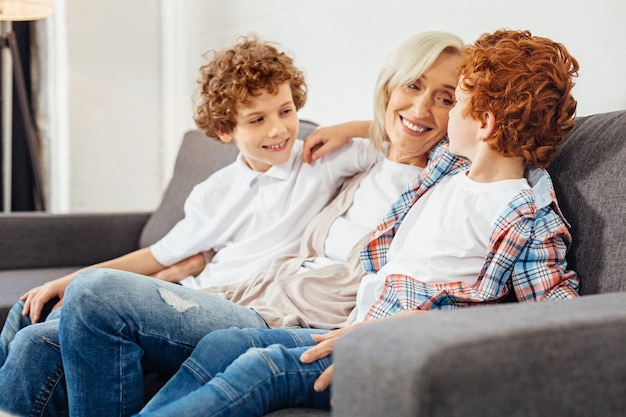 Harmonious family relations. Selective focus on a redhead little boy sitting next to his positive minded granny and his older brother while embracing on a sofa and chatting together.