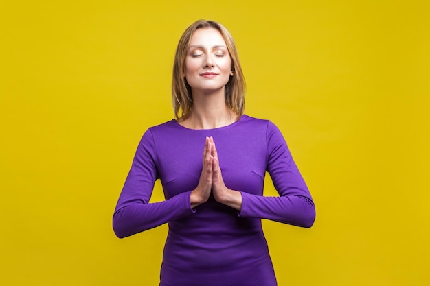 Harmony of mind. Portrait of elegant woman in purple dress standing with closed eyes and peaceful calm face meditating, holding hands in prayer. indoor studio shot isolated on yellow background