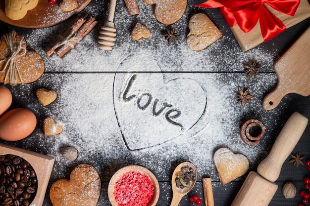 Hart getekend op bloem met de inscriptie love. peperkoekkoekjes, kruiden, koffiebonen en bakselbenodigdheden op zwarte houten achtergrond