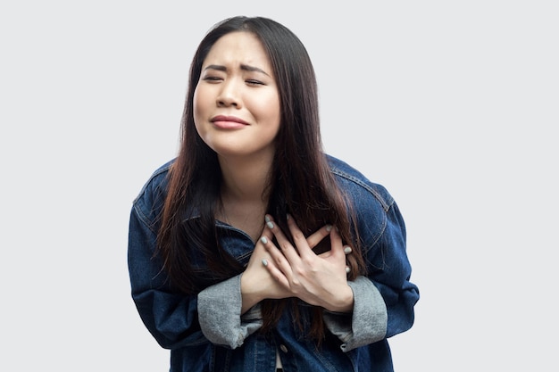Hartaanval. Portret van een bezorgde brunette aziatische jonge vrouw in een casual blauw spijkerjasje met make-up die staat en pijn op haar hart voelt. indoor studio opname, geïsoleerd op lichtgrijze achtergrond.