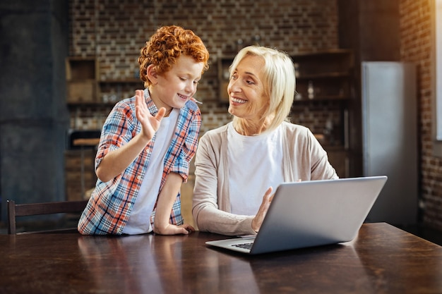 Hartverwarmende verhalen. Emotionele roodharige jongen gebaart terwijl hij een aangenaam gesprek heeft met zijn glimlachende oma die thuis op laptop werkt.