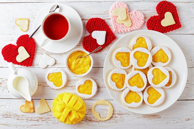Hartvormige linzer koekjes gevuld met mango jam op een witte plaat op een houten tafel versierd met rode en roze gebreide hart servetten. een kopje thee, mango en een kan met room op tafel, flatlay