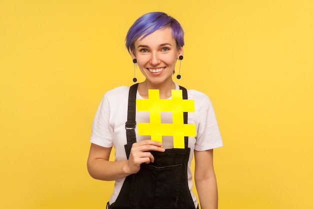 Hashtag, internetpopulariteit. Portret van een vrolijk, stijlvol hipstermeisje met violet kort haar in denimoveralls met een groot geel hekje en glimlachen. geïsoleerd op gele achtergrond, studio-opname