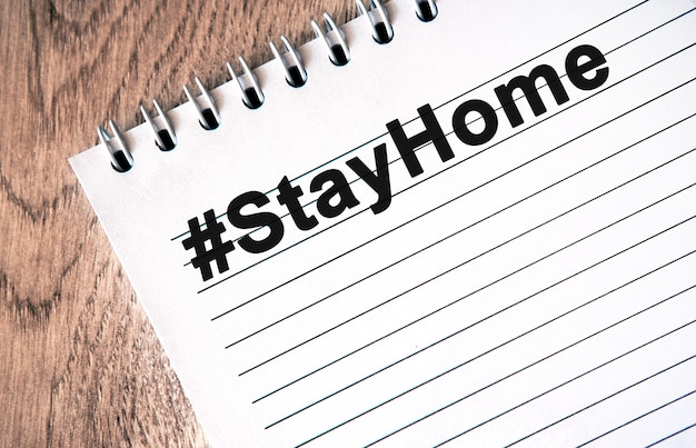 Hashtag STAYHOME - black text on a white notebook with lines on a wooden table