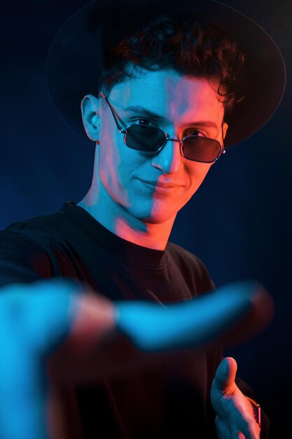 In hat and glasses Neon lighting Young european man is in the dark studio