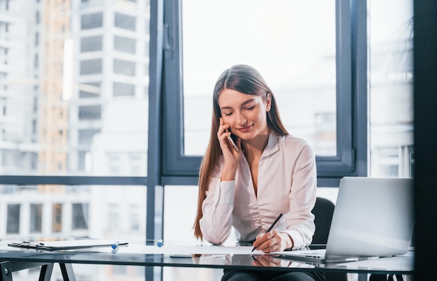 Have conversation by the phone Young woman in white formal clothes is indoors in the modern office