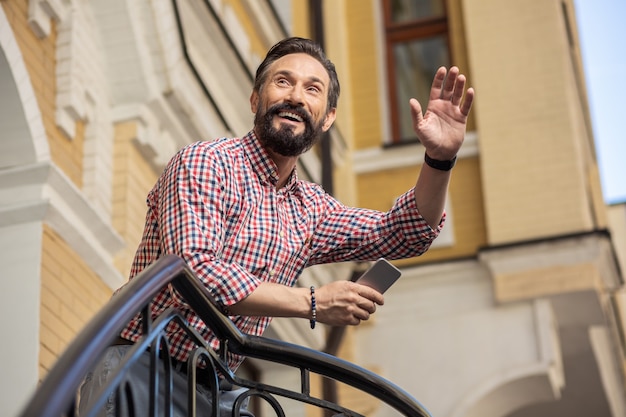 Buona giornata. basso angolo di gioioso uomo barbuto in piedi sotto il portico e saluta il suo vicino