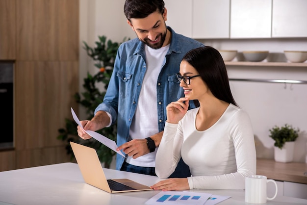 Have a look Cheerful woman working on the laptop while her young husband standing nearby