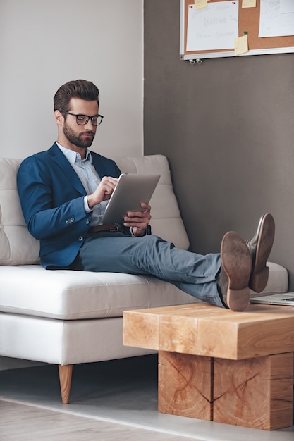 Have some work to do. Handsome young man keeping legs on table and working with touchpad while sitting on the couch in office