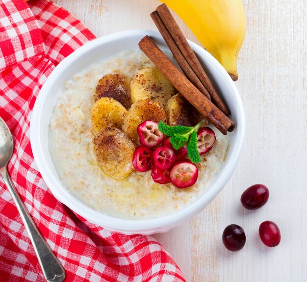 Havermoutpap met gebakken bananen, kaneel, sinaasappel en veenbessen in een keramische kom voor gezond ontbijt op een lichte achtergrond. bovenaanzicht. selectieve aandacht.