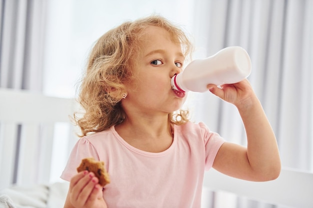 Photo having a breakfast by eating cookies cute little girl in casual clothes is indoors at home at daytime