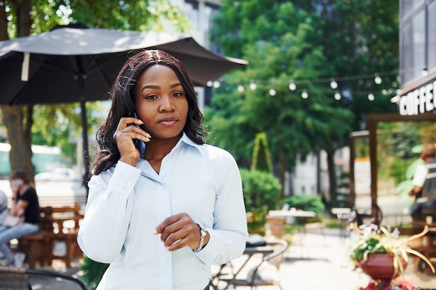 Having conversation by using phone Young afro american woman in white shirt outdoors in the city near green trees and against business building