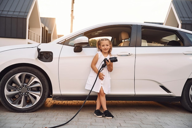 楽しい時を過す充電ケーブルを手に電気自動車の近くに立っている小さな女の子