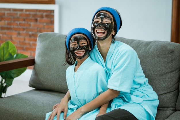 Having fun mother and daughter using facial clay masks together while relaxing on the sofa