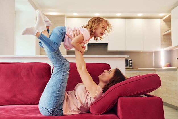 Having fun on red sofa Young mother with her little daughter in casual clothes together indoors at home