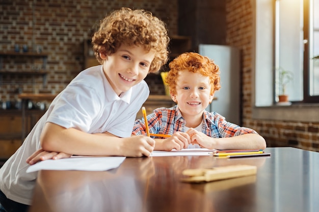 Having fun together. Selective focus on a redhead boy looking into the camera and beaming with an excitement while spending time with his older brother and drawing