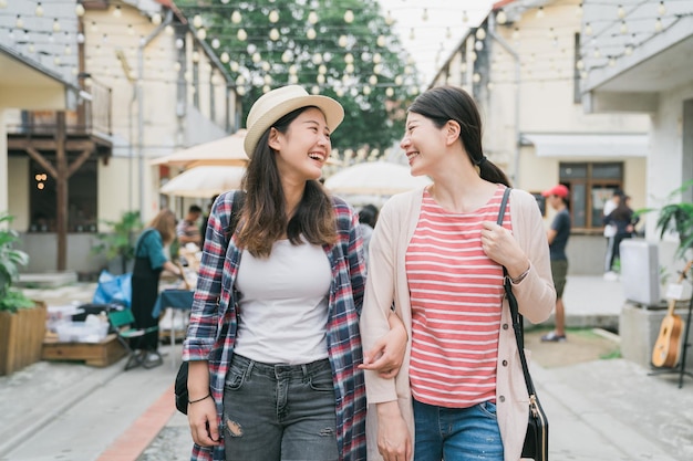 Having fun together on summer holidays. two young cheerful asian korean girls walking enjoying looking at each other and laughing on street little village. sisters relax in creative market thailand