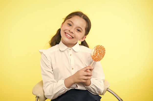 Having fun with candy Girl cute kid ponytails hairstyle eat sweet lollipop Sweets in appropriate portions ok Girl pupil school uniform likes sweet lollipop yellow background Her favorite flavour