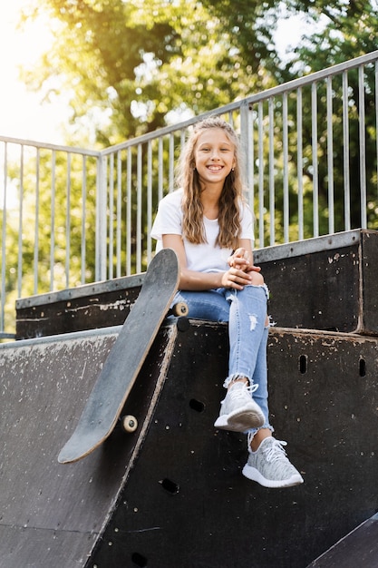 スケートボードを楽しんでいるスポーツランプに座っているスケートの面白い子の女の子は、スケートの遊び場で笑顔と顔をゆがめますスケートボードでポーズをとるアクティブなティーンエイジャー極端なライフスタイル
