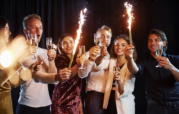 Having fun with sparklers. Group of cheerful friends celebrating new year indoors with drinks in hands.