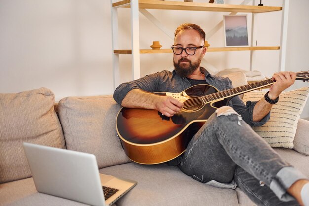 Photo having good time practicing man in casual clothes and with acoustic guitar is indoors