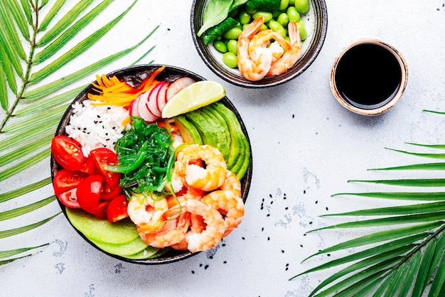 Hawaiiaanse poke bowl met garnalen avocado radijs wortel tomaat chuka zeewier en witte rijst Sojasaus limoen en sesam dressing Witte tafel achtergrond palmbladeren bovenaanzicht