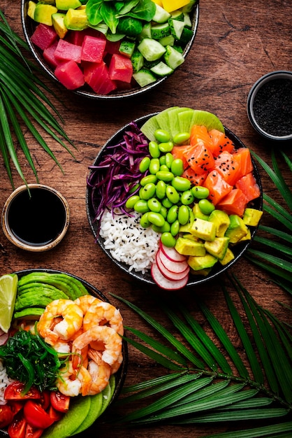 Hawaiiaanse poke bowl set tonijn zalm garnalen met avocado mango radijs rijst en andere ingrediënten Sojasaus en sesam dressing Houten tafel achtergrond palmbladeren bovenaanzicht