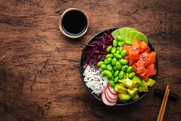 Hawaiian poke bowl with salmon avocado radish edamame beans red cabbage and white rice Soy sauce lime and sesame dressing Old wood table background palm leaves top view