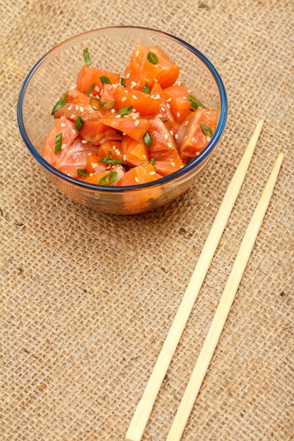 Hawaiian salmon poke with green onions and sesame seeds in glass bowl with chopsticks on sackcloth