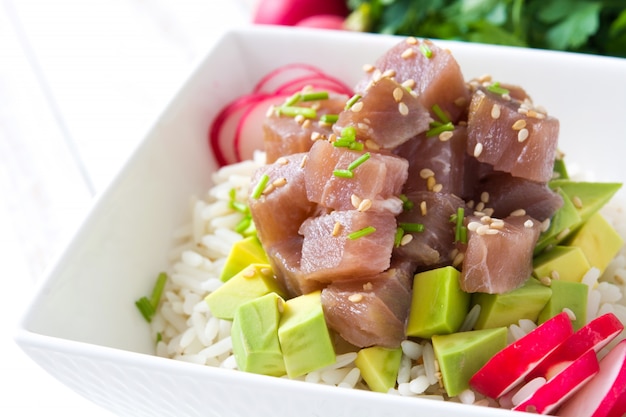 Hawaiian tuna poke bowl with avocado, radishes and sesame seeds on white wooden table close up