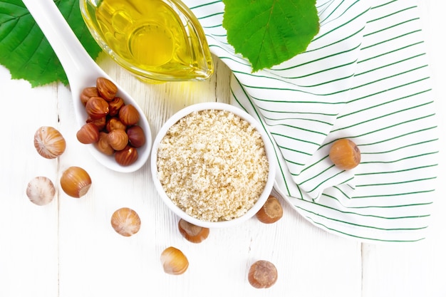 Hazelnut flour in a bowl, oil in a glass gravy boat, nuts, napkin and filbert branch with green leaves on wooden board background from above