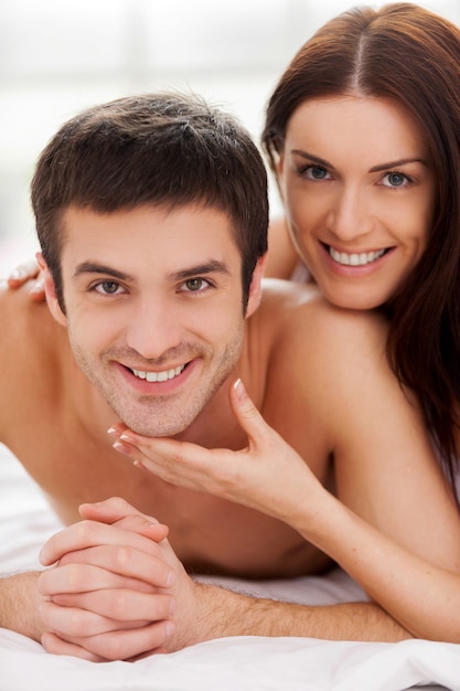 He is mine! Cheerful young loving couple lying in bed and smiling while woman touching her boyfriend chin