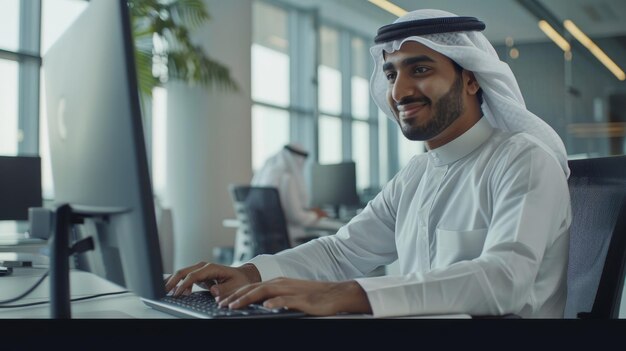 He is a smiling young middle eastern businessman sitting at a desk in a modern office typing on his computer keyboard sending emails to his business partners and doing marketing research