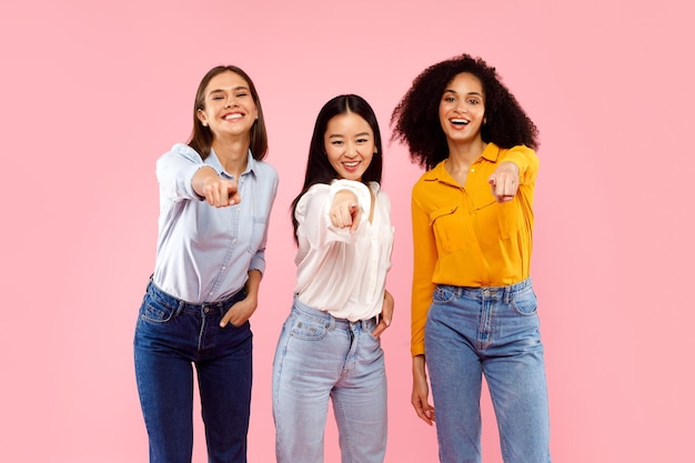 Hé jij portret van gelukkige multi-etnische dames die met de vingers naar de camera wijzen en lachend poseren over roze