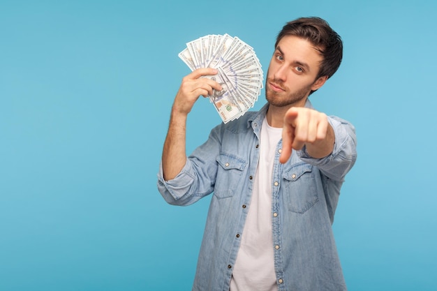 Hé jij, tijd om geld te verdienen. Portret van een knappe man in een spijkerblouse die naar de camera wijst en dollarbankbiljetten laat zien, die aanmoedigt om winst te maken, de loterij te winnen. studio-opname geïsoleerd op blauwe achtergrond