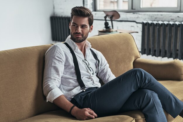 He knows all of your secrets. Handsome young man in white shirt and suspenders looking away and smiling while sitting on sofa