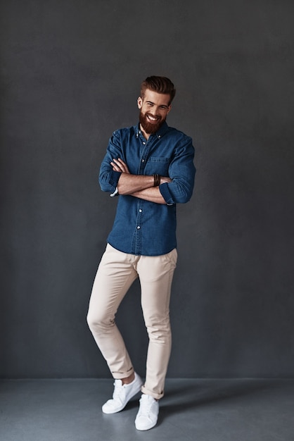 He knows everything about you. Full length of handsome young man looking at camera and keeping arms crossed while standing against grey background